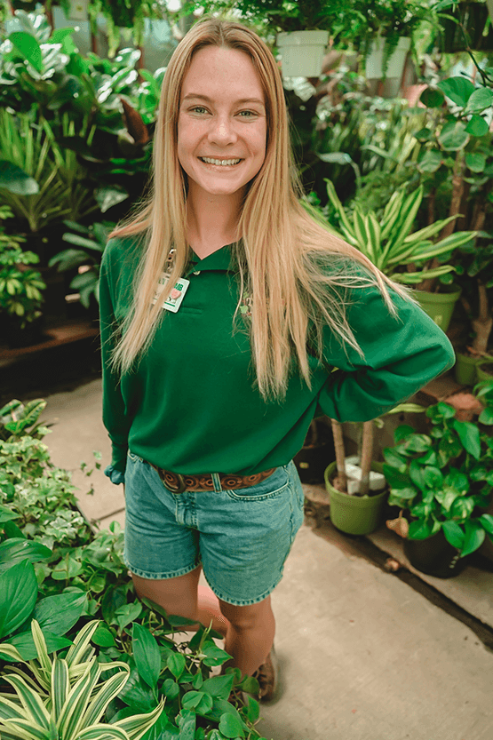 In a greenhouse full of plants, a young woman in a green shirt tends to the best garden center near me.