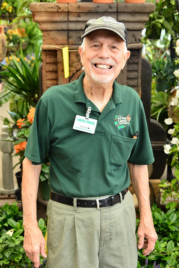 Smiling elderly man wearing a green nursery uniform and a cap stands in front of plants and garden decor, with a nametag reading “Green Thumb.”.
