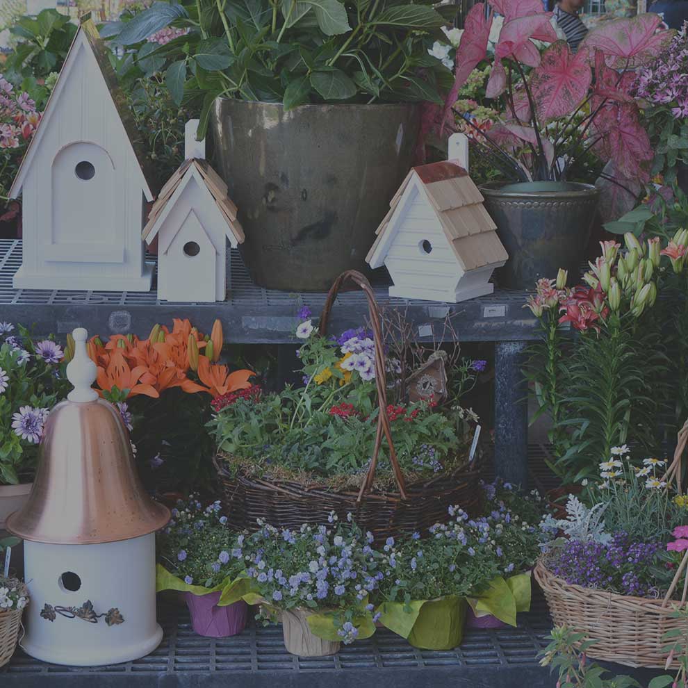 A variety of birdhouses, potted flowers, and baskets of plants are displayed on metal shelves at a garden shop.