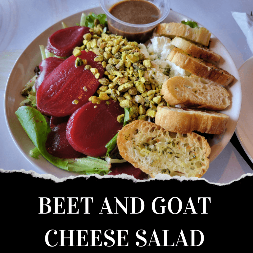 A plate of beet and goat cheese salad with mixed greens, sliced beets, crumbled goat cheese, chopped nuts, baguette slices, and a side of vinaigrette dressing.