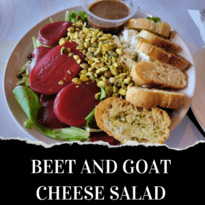 A plate of beet and goat cheese salad with mixed greens, sliced beets, crumbled goat cheese, chopped nuts, baguette slices, and a side of vinaigrette dressing.