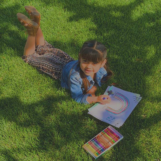 A young girl lies on grass drawing a rainbow with markers in a sketchbook; a box of colorful markers is beside her.