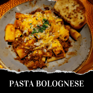 A plate of rigatoni pasta with meat sauce, grated cheese, chopped herbs, and a slice of garlic bread; text reads "Pasta Bolognese.