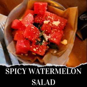 A bowl of spicy watermelon salad with cubed watermelon, herbs, and crumbled cheese, served on brown parchment paper.