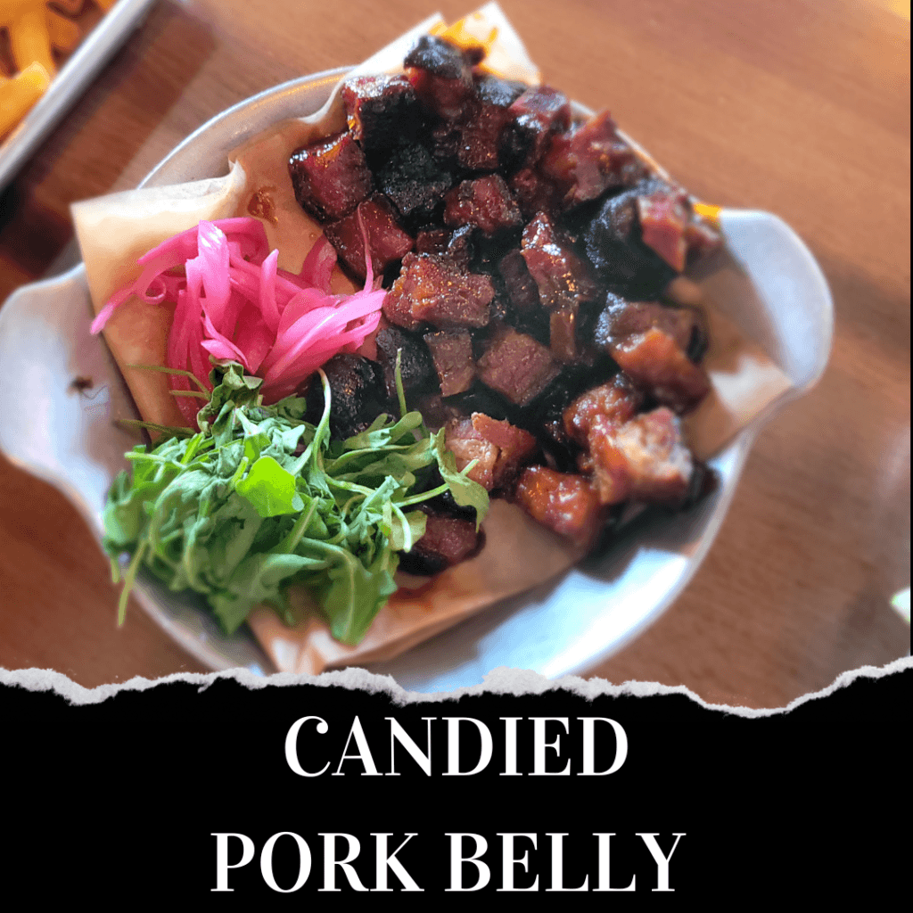 A bowl of candied pork belly cubes served with pickled onions and fresh greens on parchment paper.