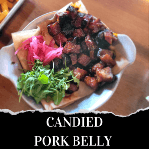 A bowl of candied pork belly cubes served with pickled onions and fresh greens on parchment paper.