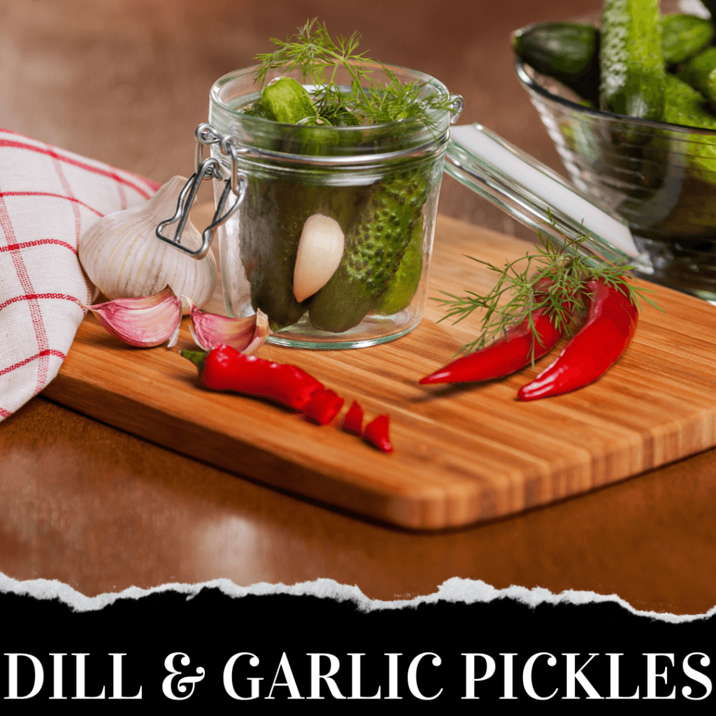 Glass jar of pickles with garlic, dill, and red chili peppers on a wooden cutting board, next to a bowl of cucumbers and a red and white cloth. Text reads "Dill & Garlic Pickles.