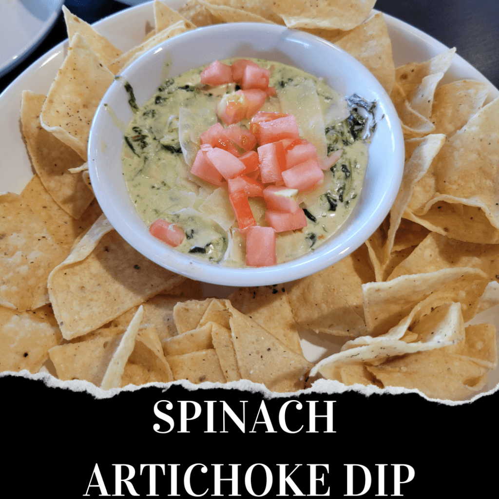 A bowl of spinach artichoke dip topped with chopped tomatoes, surrounded by tortilla chips on a white plate. The words "Spinach Artichoke Dip" appear at the bottom.