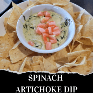 A bowl of spinach artichoke dip topped with chopped tomatoes, surrounded by tortilla chips on a white plate. The words "Spinach Artichoke Dip" appear at the bottom.