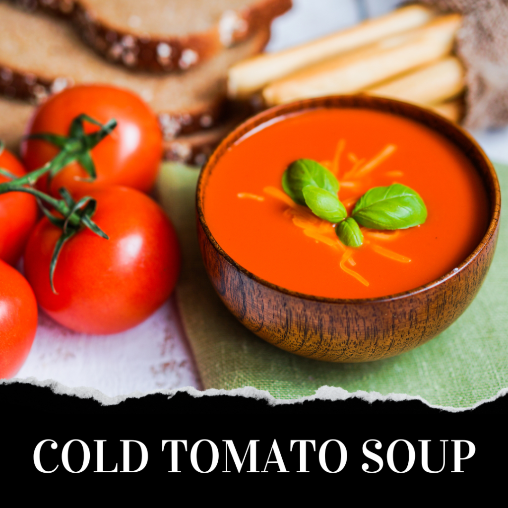 A wooden bowl of cold tomato soup garnished with basil, next to fresh tomatoes, bread slices, and breadsticks.