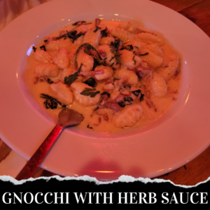 A plate of gnocchi in a creamy herb sauce, topped with chopped herbs. A spoon is resting on the plate. The text at the bottom reads "GNOCCHI WITH HERB SAUCE.
