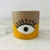 A small cylindrical pot with a beige and yellow design featuring a stylized eye in the middle.