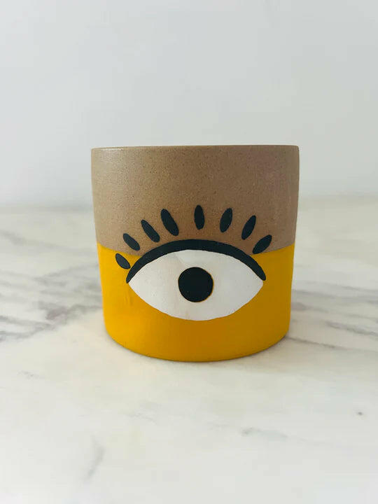 A small cylindrical pot with a beige and yellow design featuring a stylized eye in the middle.