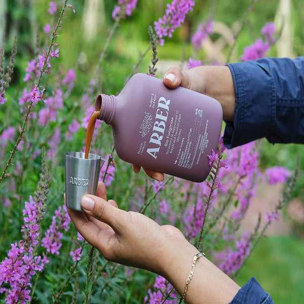 A person pours a liquid from a mauve ARBER bottle into a metal cup, with purple flowers and greenery in the background.