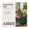 Packaging for an Arber Repotting Mat, showing product features and an hands repotting a plant. Box includes a small circular cutout displaying the mat’s color.